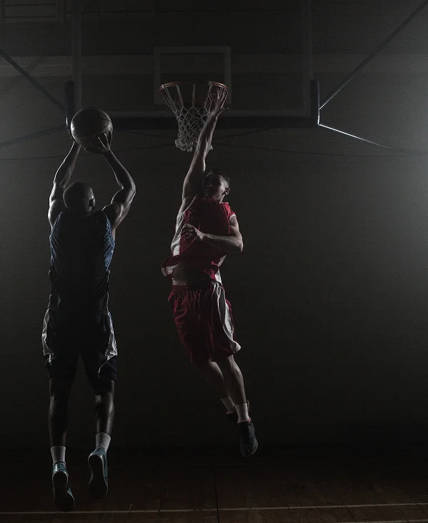 Dois jogadores de basquetebol saltam perto do cesto numa ginásio pouco iluminado. Um de vermelho tenta marcar, enquanto o outro de preto tenta bloquear. A cena é intensa e dramática.